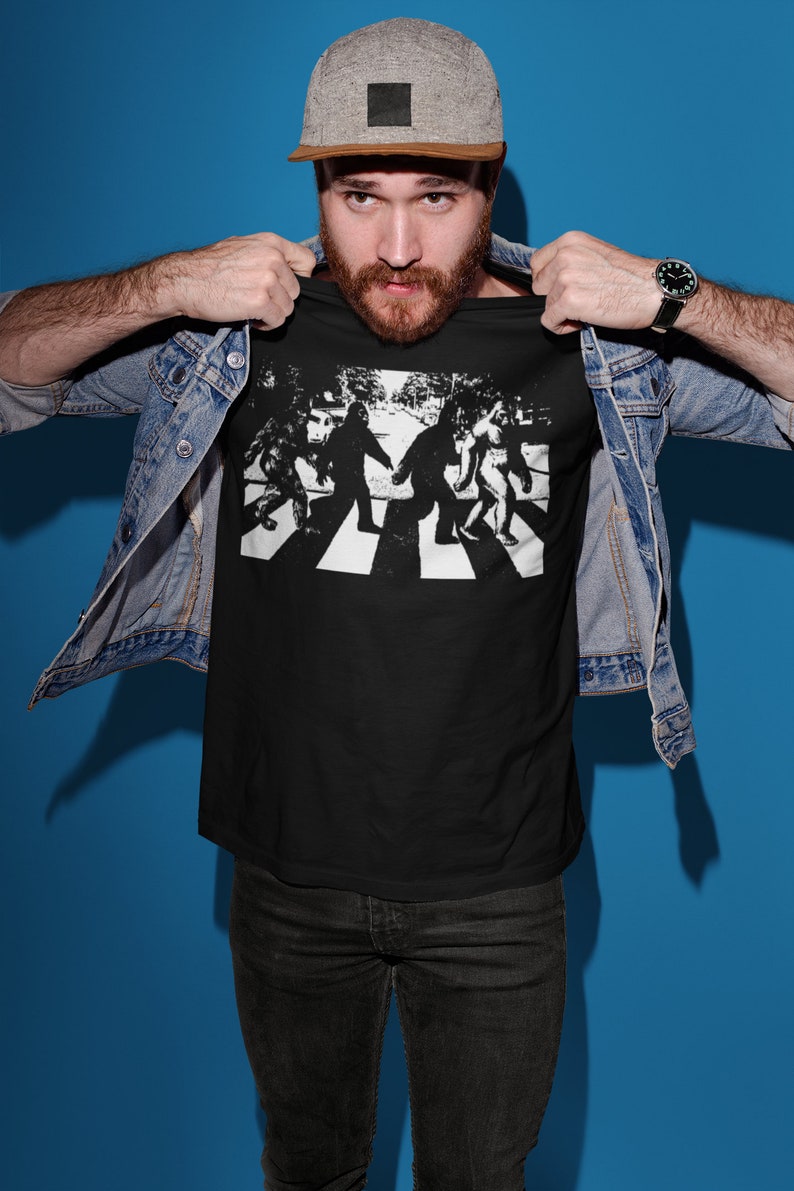 Puede incluir: Una camiseta negra con un gr&aacute;fico blanco de cuatro figuras de Pie Grande caminando por un paso de cebra, similar a la ic&oacute;nica portada del &aacute;lbum de los Beatles.