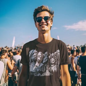Puede incluir: Camiseta gris oscuro con un estampado gr&aacute;fico de tres figuras, incluyendo un gorila y una criatura parecida a una polilla. La persona lleva gafas de sol y sonr&iacute;e. El fondo es una multitud de personas bajo un cielo azul.