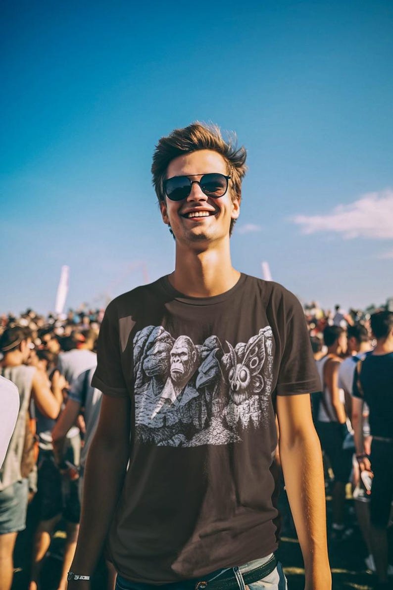 Puede incluir: Camiseta gris oscuro con un estampado gr&aacute;fico de tres figuras, incluyendo un gorila y una criatura parecida a una polilla. La persona lleva gafas de sol y sonr&iacute;e. El fondo es una multitud de personas bajo un cielo azul.