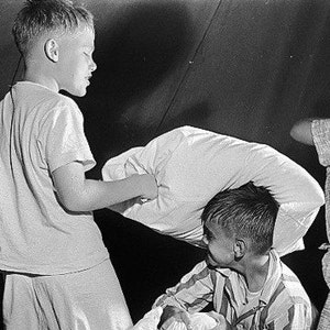 May include: Four boys are playing a pillow fight in a tent. They are all wearing white shirts and shorts. One boy is holding a pillow over his head, while another boy is swinging a pillow at him. The other two boys are watching the action.