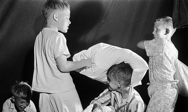 May include: Four boys are playing a pillow fight in a tent. They are all wearing white shirts and shorts. One boy is holding a pillow over his head, while another boy is swinging a pillow at him. The other two boys are watching the action.