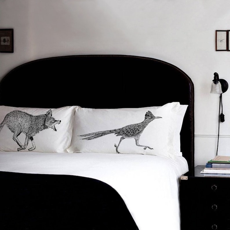 May include: Two white pillowcases with black and white illustrations of a coyote and a roadrunner. The pillowcases are on a black headboard bed.