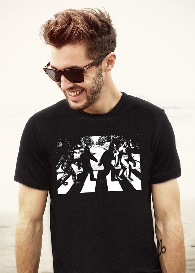 Puede incluir: Una camiseta negra con un gr&aacute;fico blanco de un grupo de figuras de Bigfoot caminando por un paso de cebra, similar a la portada del &aacute;lbum Abbey Road de los Beatles.