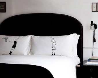 May include: Two white pillowcases featuring black line drawings of basketball players. One shows a player shooting a basket, the other a free throw. The pillowcases are on a bed with a black headboard. The bed has a black blanket.