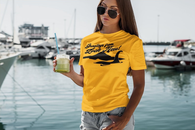 Może przedstawiać: Kobieta w ż&oacute;łtej koszulce z czarnym nadrukiem "Greetings from Loch Ness!" i czarną sylwetką potwora z Loch Ness. Trzyma zielony smoothie w szklanym słoiku ze słomką.