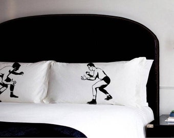 May include: White pillowcases with black illustrations of wrestlers in action. The pillows are on a white bed with a black headboard. A black nightstand and wall sconce are visible in the background.