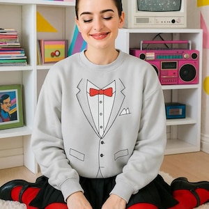 May include: A light gray sweatshirt with a tuxedo design, featuring a black outline of a suit jacket, a red bow tie, and a white shirt. The person is wearing a black skirt, red tights, and black shoes. The background includes a white shelf with a retro TV and a pink boombox.