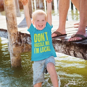 May include: A young boy wearing a blue t-shirt with the text "Don't hassle me I'm local" in yellow letters is being held up by two adults on a wooden dock over a body of water. The boy is smiling and has blonde hair.