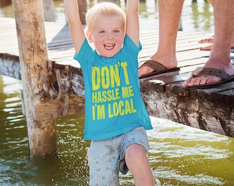 Könnte beinhalten: Ein kleiner Junge trägt ein blaues T-Shirt mit dem Text "Don't hassle me I'm local" in gelben Buchstaben. Er wird von zwei Erwachsenen auf einem Holzsteg über einem Gewässer hochgehalten. Der Junge lächelt und hat blonde Haare.
