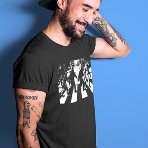 Puede incluir: Una camiseta negra con un gr&aacute;fico blanco de cuatro figuras caminando por un paso de cebra. Las figuras est&aacute;n estilizadas para parecerse a Bigfoot.