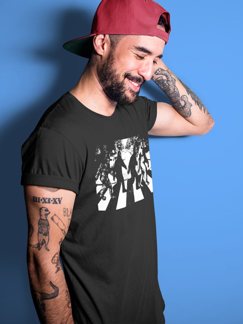 Puede incluir: Una camiseta negra con un gr&aacute;fico blanco de cuatro figuras caminando por un paso de cebra. Las figuras est&aacute;n estilizadas para parecerse a Bigfoot.