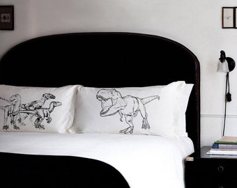 Könnte beinhalten: Weiße Kissenbezüge mit schwarzen Dinosaurier-Illustrationen. Einer zeigt einen Tyrannosaurus Rex, der andere eine Gruppe von Dinosauriern. Die Bettwäsche befindet sich auf einem Bett mit schwarzem Kopfteil und schwarzer Tagesdecke.