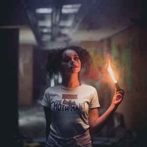 May include: A woman wearing a white t-shirt with the text "Beware the Mothman!" and a black bat graphic. She is holding a lit flare in a dark, industrial setting.