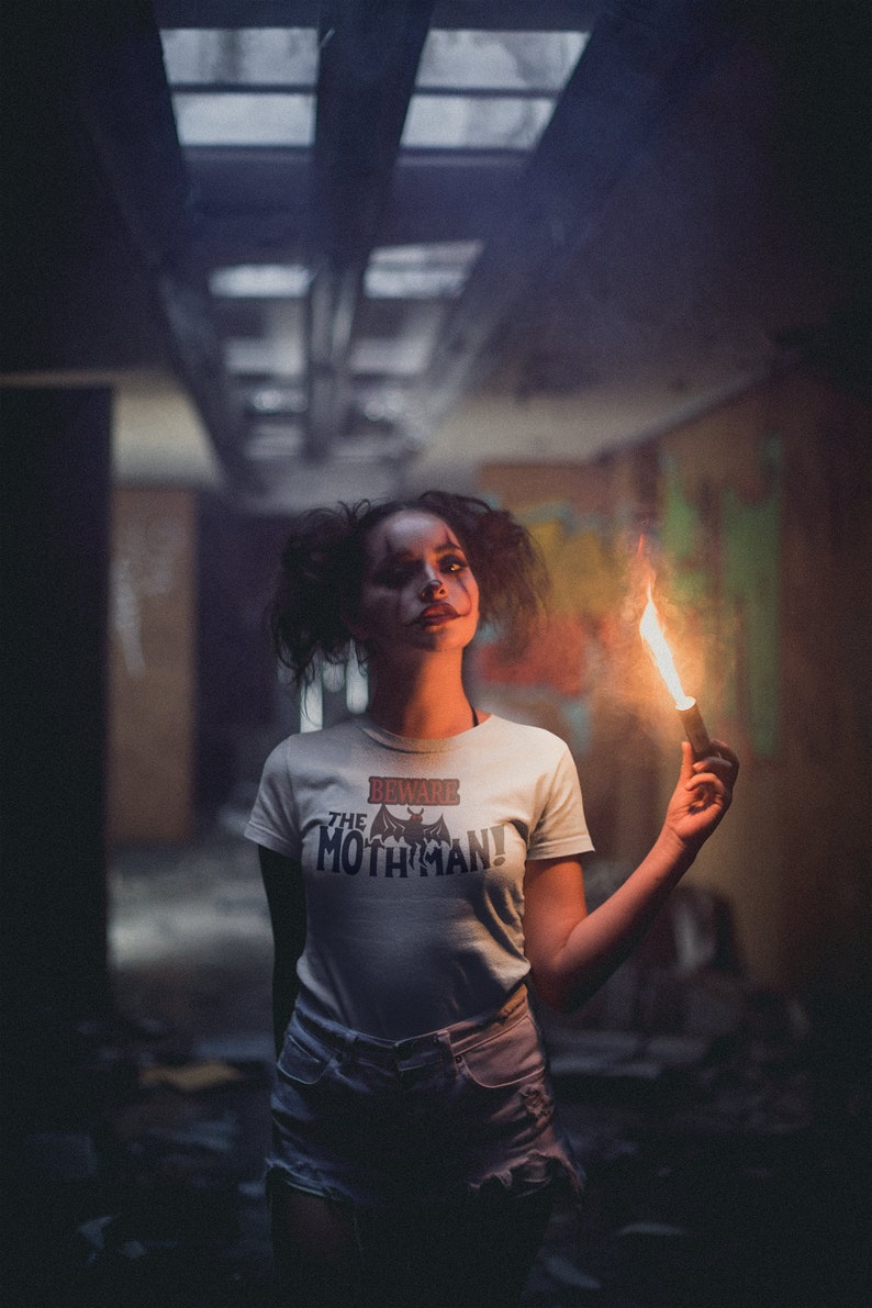 May include: A woman wearing a white t-shirt with the text "Beware the Mothman!" and a black bat graphic. She is holding a lit flare in a dark, industrial setting.