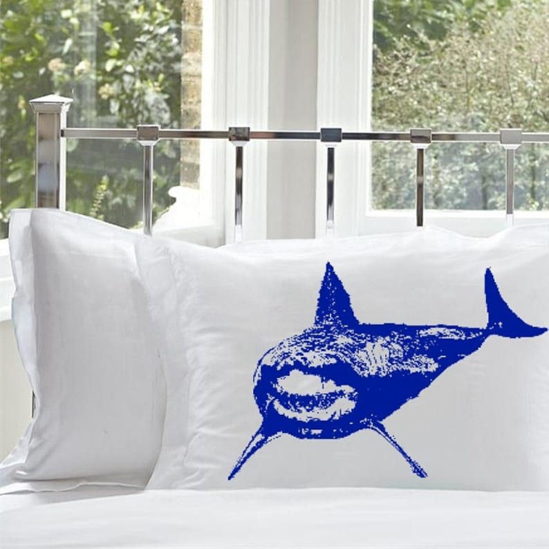 May include: Two white pillowcases with a blue shark silhouette printed on one of the pillowcases.