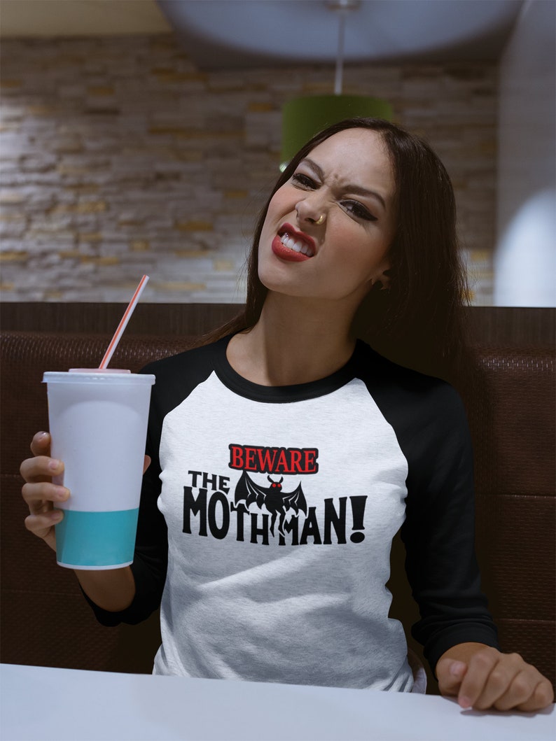 May include: A white and black baseball style t-shirt with a black bat graphic and the text "Beware the Mothman!" in red and black lettering.