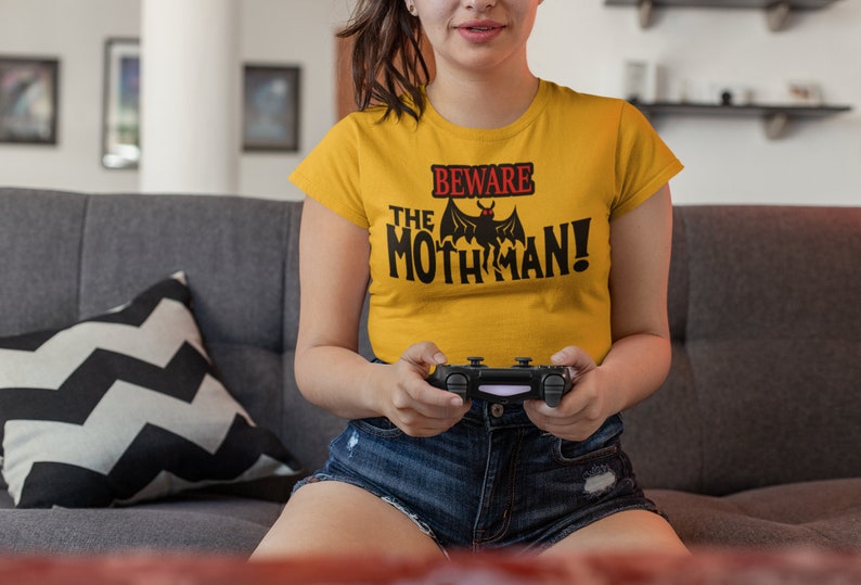 May include: A woman wearing a yellow t-shirt with the text "Beware the Mothman!" and a black bat graphic. She is holding a black video game controller.