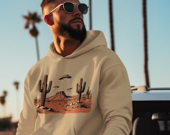 May include: A beige hoodie with a desert landscape graphic featuring cacti and UFOs. The graphic is in shades of brown, orange, and grey. The hoodie has a front pocket and a drawstring hood. The background shows palm trees and a blue sky.