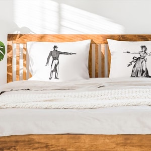May include: Two white pillowcases with black and white illustrations of people. The pillowcases are on a wooden bed frame with a white blanket.