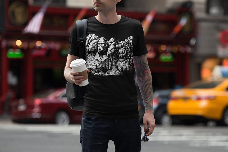 Puede incluir: Camiseta negra de cuello redondo con un gr&aacute;fico blanco que representa un gorila, un humano y una criatura parecida a una polilla, una parodia del Monte Rushmore. La persona lleva vaqueros azules y un vaso de caf&eacute;.