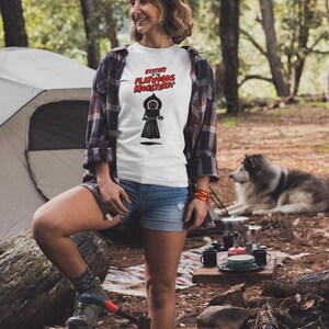 May include: White t-shirt with the text "Mystery of the Flatwoods Monster" in red and black. The shirt features a graphic of a black figure with a glowing red eye. The person is wearing denim shorts and a plaid shirt.