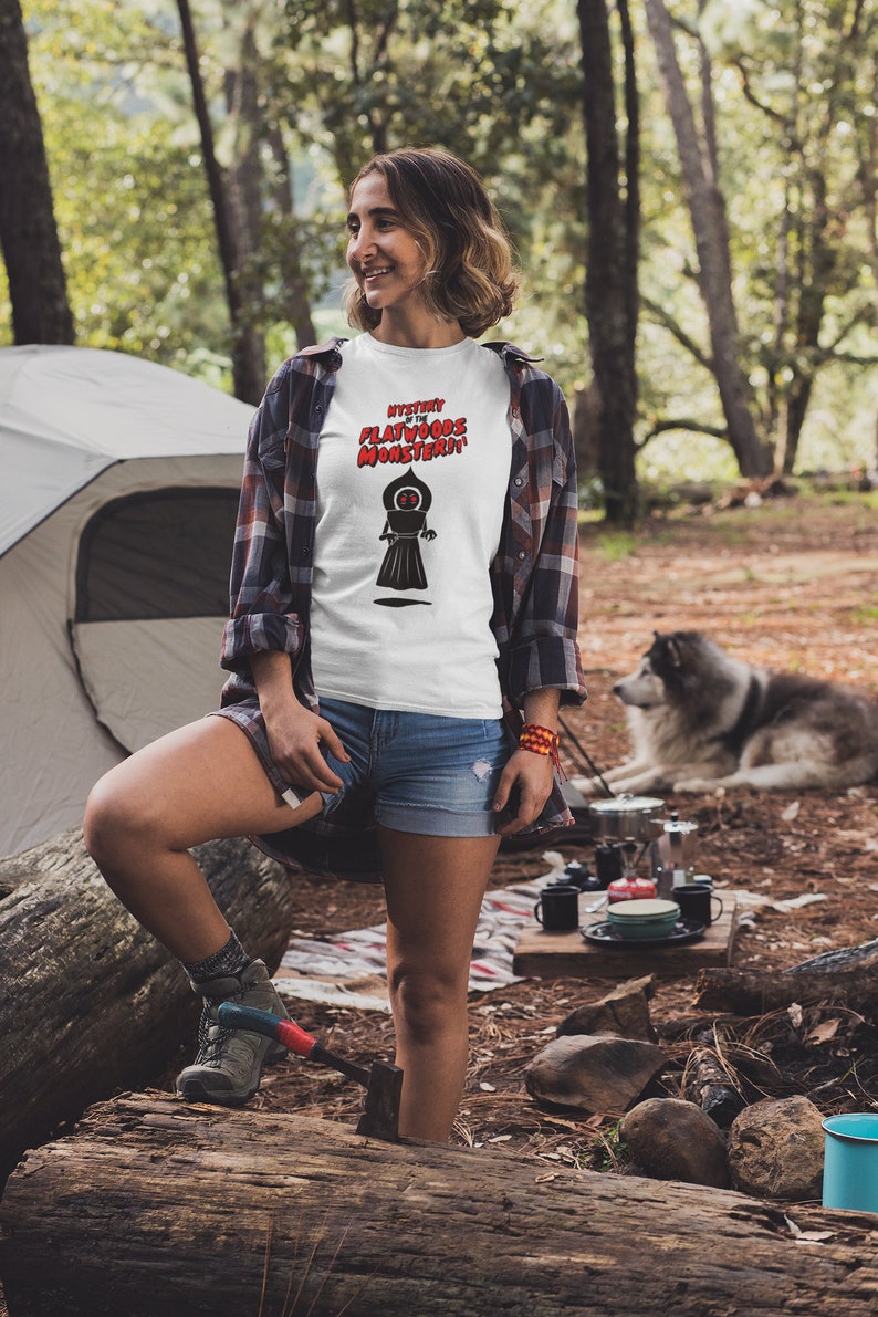 May include: White t-shirt with the text "Mystery of the Flatwoods Monster" in red and black. The shirt features a graphic of a black figure with a glowing red eye. The person is wearing denim shorts and a plaid shirt.