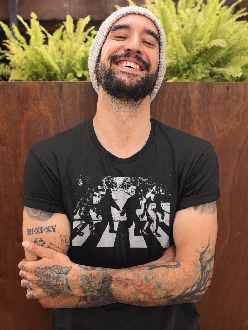 Puede incluir: Una camiseta negra con un gr&aacute;fico de cuatro figuras caminando por un paso de cebra, similar a la portada del &aacute;lbum Abbey Road de los Beatles. Las figuras est&aacute;n estilizadas para parecerse a Bigfoot.