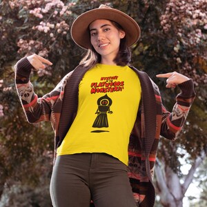 May include: Yellow t-shirt with the text "Mystery of the Flatwoods Monster!!" in red and black. The shirt features a black graphic of a cloaked figure. The person is wearing a brown hat, a patterned cardigan, and olive green pants.