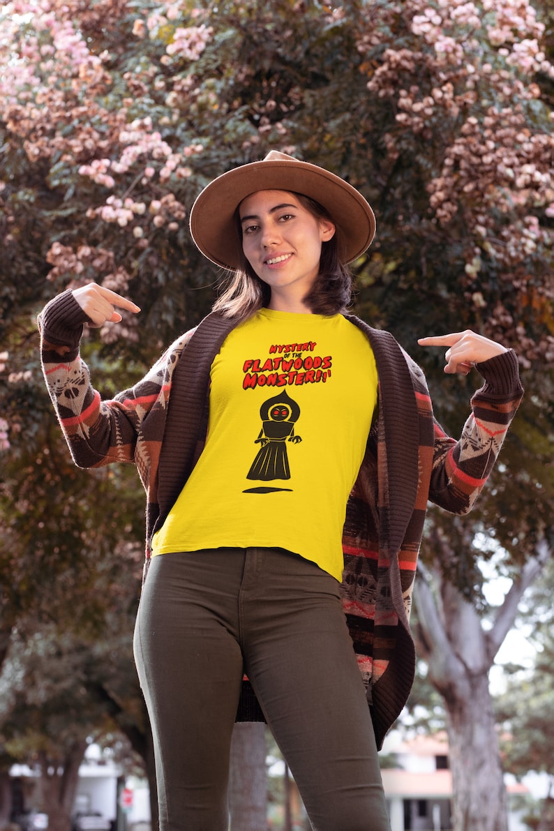 May include: Yellow t-shirt with the text "Mystery of the Flatwoods Monster!!" in red and black. The shirt features a black graphic of a cloaked figure. The person is wearing a brown hat, a patterned cardigan, and olive green pants.