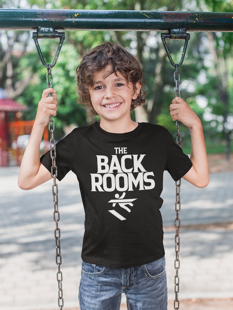 May include: A black t-shirt with white text that reads "THE BACK ROOMS" and a white graphic of a person falling down stairs. The shirt is being worn by a child on a swing set.