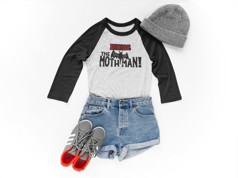 May include: A gray and black baseball tee shirt with the text "Beware the Mothman!" in red and black. The shirt is paired with blue denim shorts, gray sneakers, and a gray beanie.
