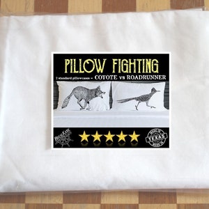 May include: A set of two standard pillowcases featuring a black and white illustration of a coyote chasing a roadrunner. The pillowcases are packaged in a white plastic bag. The text "Pillow Fighting - Coyote vs Roadrunner" is printed on the packaging. The packaging also includes five gold stars and a "Made in Texas" stamp.