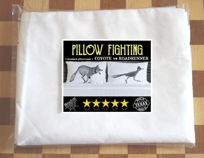 May include: A set of two standard pillowcases featuring a black and white illustration of a coyote chasing a roadrunner. The pillowcases are packaged in a white plastic bag. The text "Pillow Fighting - Coyote vs Roadrunner" is printed on the packaging. The packaging also includes five gold stars and a "Made in Texas" stamp.