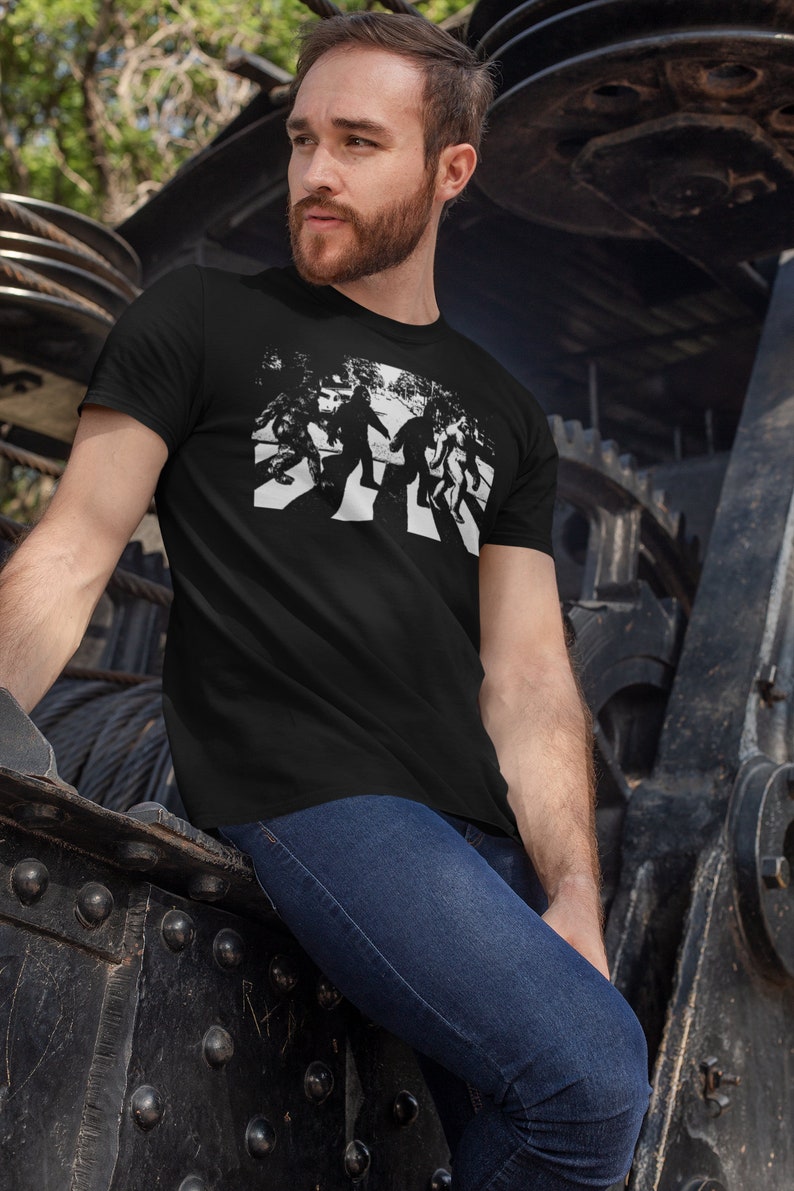 Puede incluir: Una camiseta negra con un gr&aacute;fico blanco de cuatro figuras caminando por un paso de cebra. Las figuras est&aacute;n estilizadas para parecerse a Bigfoot.