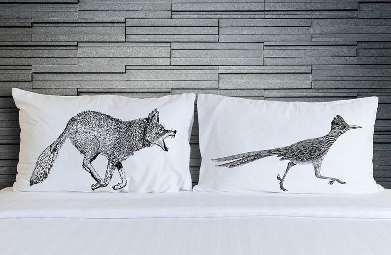 May include: Two white pillowcases with black and white illustrations of a coyote and a roadrunner. The coyote is running and has its mouth open, while the roadrunner is running with its wings spread out.