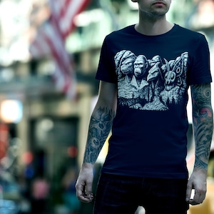 Puede incluir: Camiseta azul marino con un dise&ntilde;o gr&aacute;fico en blanco y negro que parodia el Monte Rushmore con criaturas. La camiseta es de cuello redondo y parece estar hecha de un material suave. El fondo es una escena urbana borrosa.