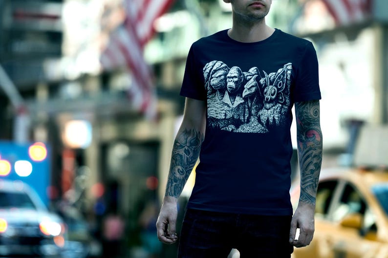 Puede incluir: Camiseta azul marino con un dise&ntilde;o gr&aacute;fico en blanco y negro que parodia el Monte Rushmore con criaturas. La camiseta es de cuello redondo y parece estar hecha de un material suave. El fondo es una escena urbana borrosa.