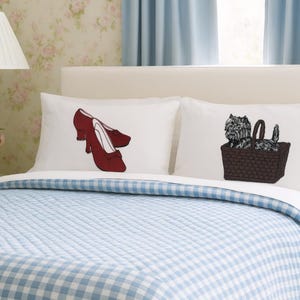 May include: Two white pillowcases on a bed, each with a different illustration. One pillow features a pair of red shoes, and the other shows a dog in a basket. The bed has a blue and white checkered quilt.