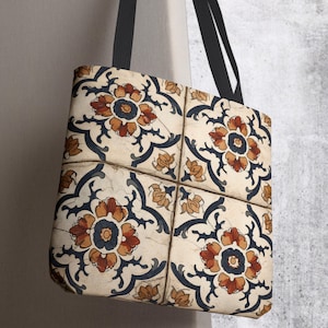 May include: A square tote bag featuring a repeating pattern of four large floral motifs in shades of burnt orange, dark blue, and cream. The design mimics the appearance of ceramic tiles with thin lines separating the sections. It has black straps.