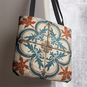 May include: A tote bag featuring a repeating tile pattern in shades of cream, teal, and burnt orange. The design displays a symmetrical floral motif with distressed, weathered textures across the surface and two black fabric shoulder straps.