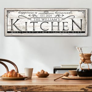 May include: A rectangular wooden sign with the text "happiness is homemade" and "THE WILLIAM'S KITCHEN" in black lettering. The sign also includes the text "MEALS AND MEMORIES MADE HERE" and "EST. 2024". The sign is hanging on a white wall.