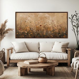 May include: A framed canvas art piece depicting a field of wildflowers in shades of brown, yellow, and red. The artwork hangs above a light-colored sofa with white cushions. A wooden coffee table with a teapot and books sits in front of the sofa.