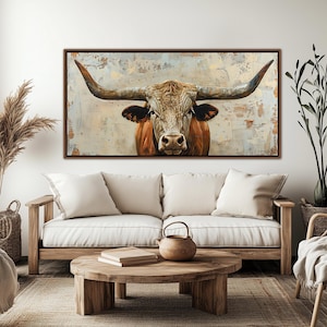 May include: A framed canvas print depicting a longhorn bull with large horns and a brown and tan coat. The background features a textured, neutral colour palette. The artwork is displayed above a sofa in a living room setting.