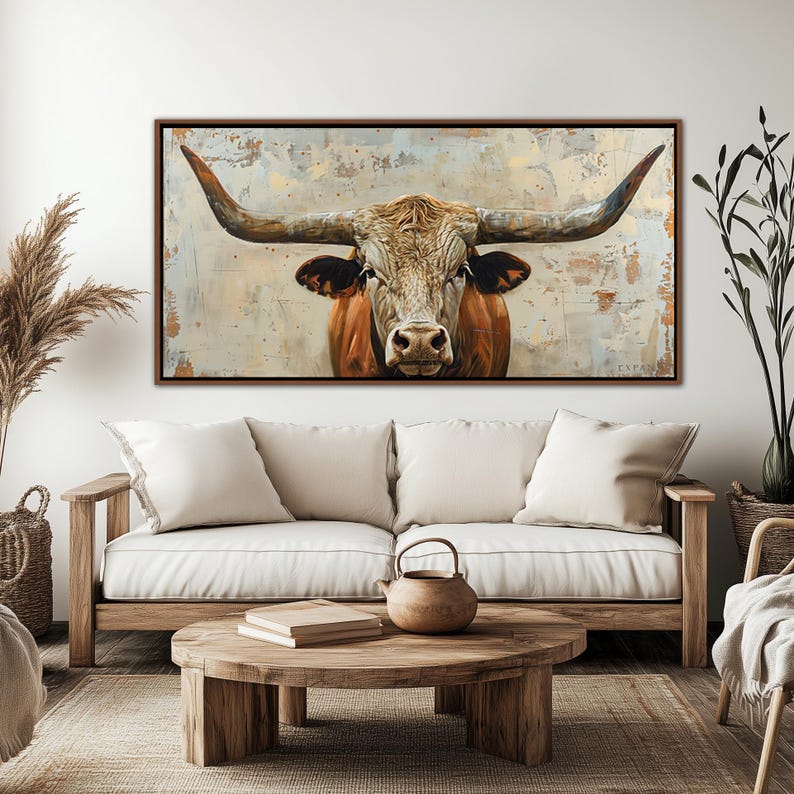 May include: A framed canvas print depicting a longhorn bull with large horns and a brown and tan coat. The background features a textured, neutral colour palette. The artwork is displayed above a sofa in a living room setting.
