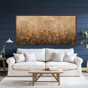 May include: A framed canvas art piece depicting a field of wildflowers in shades of brown, yellow, and red. The artwork is set against a neutral background and framed in a dark brown border. The painting hangs above a white sofa.