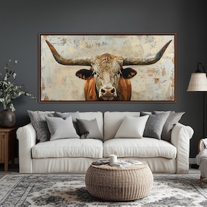 May include: A framed canvas print of a longhorn bull, with a detailed depiction of its head and horns. The artwork has a textured background in neutral tones. The print is displayed above a white sofa with grey cushions, in a living room setting.