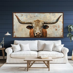 May include: A framed canvas print featuring a longhorn bull. The bull is shown head-on, with prominent horns and a brown and white coat. The background has a textured, neutral colour palette. The artwork is displayed above a white sofa.