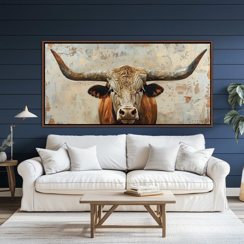 May include: A framed canvas print featuring a longhorn bull. The bull is shown head-on, with prominent horns and a brown and white coat. The background has a textured, neutral colour palette. The artwork is displayed above a white sofa.