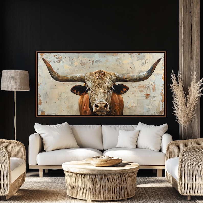 May include: A framed canvas print depicting a longhorn bull's head with large, curved horns. The bull has a brown and white coat. The artwork is displayed in a living room with a white sofa and a woven coffee table, creating a rustic aesthetic.