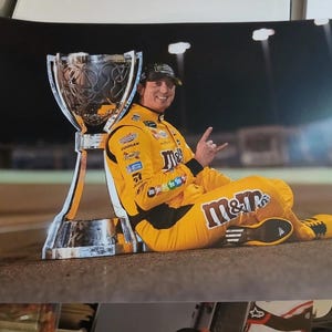 Pode incluir: Fotografia de um piloto de corridas com um fato de corrida amarelo com detalhes em preto, sentado ao lado de um grande troféu. O fato apresenta o logótipo da M&M's. O piloto usa um boné preto e faz um gesto com a mão.